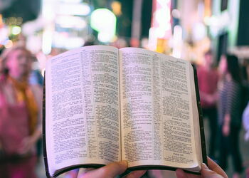 Geöffnete Bibel in den Händen einer Person vor unscharfem nächtlichem Stadtbild mit Menschen im Hintergrund; Symbolbild für den weltweiten Zugang zur Bibel in verschiedenen Sprachen.