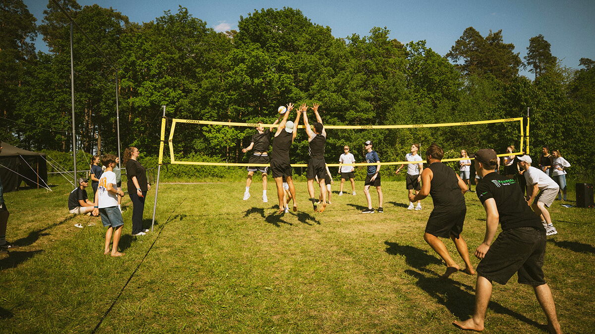 Jugendliche spielen Beachvolleyball auf einer Wiese während der ONECON 2026. Mehrere Teilnehmer stehen am Netz, weitere Zuschauer und Mitspieler befinden sich am Spielfeldrand.