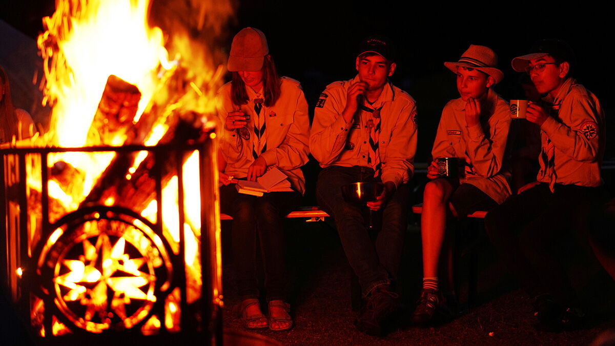 Flammen lodern aus einem Feuerkorb mit Royal Rangers Emblem, vier Menschen sitzen dahinter.
