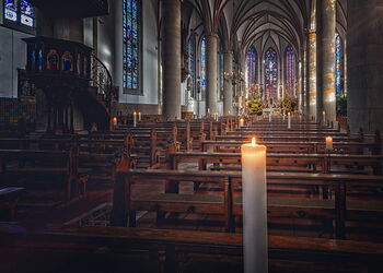 Innenraum einer großen Kirche mit hohen Gewölben, bunten Glasfenstern und Holzbänken; im Vordergrund brennt eine einzelne Kerze, der Raum wirkt ruhig und lichtdurchflutet.