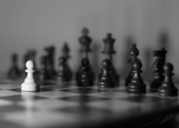 Schwarz-weißes Schachbrett mit einer einzelnen weißen Figur im Vordergrund, umgeben von unscharfen schwarzen Figuren im Hintergrund.