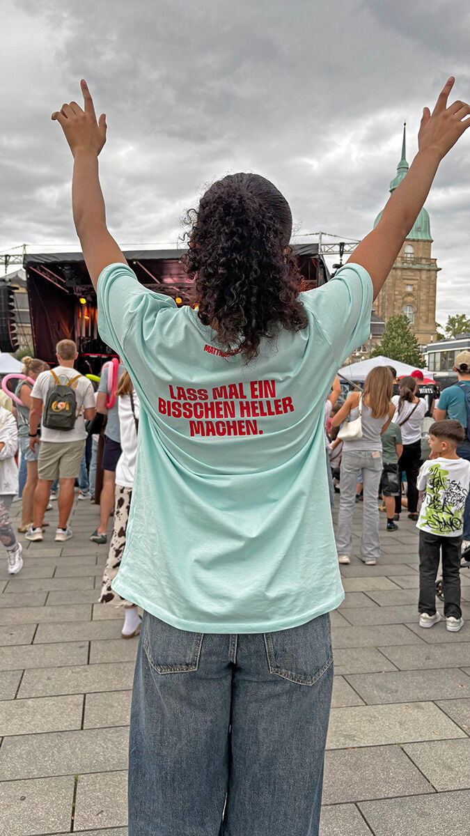 Eine junge Frau steht mit erhobenen Armen vor der Festivalbühne. Auf ihrem hellblauen HELLER-Shirt steht in roten Buchstaben: „Lass mal ein bisschen heller machen.“