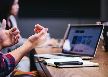 Mann vor einem aufgeklapptem Notebook mit einem Notitzbuch auf dem Tisch und einer sitzenden Frau im Hintergrund