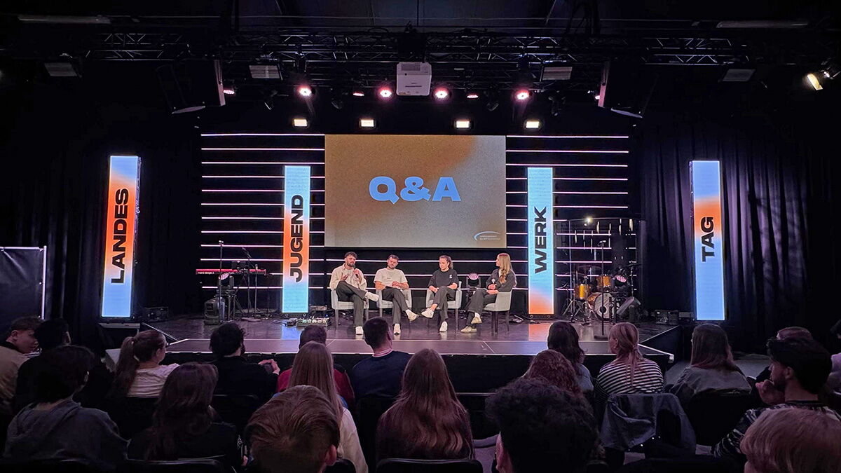 Mehrere Personen sitzen auf einer Bühne bei einem Q&A-Podium, davor Publikum; große Leinwand und beleuchtete Banner mit „Jugend“, „Werk“ und „Tag“