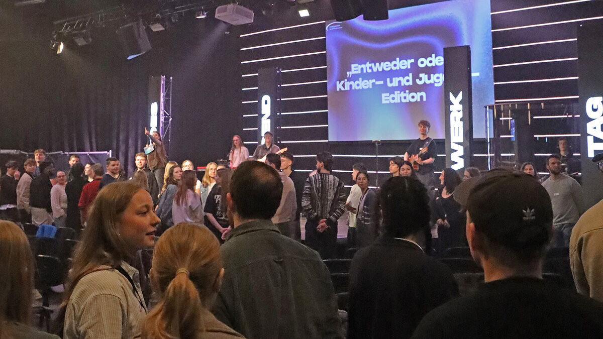 Viele junge Erwachsene stehen und unterhalten sich vor einer Bühne mit Band und Präsentation während einer Veranstaltung.