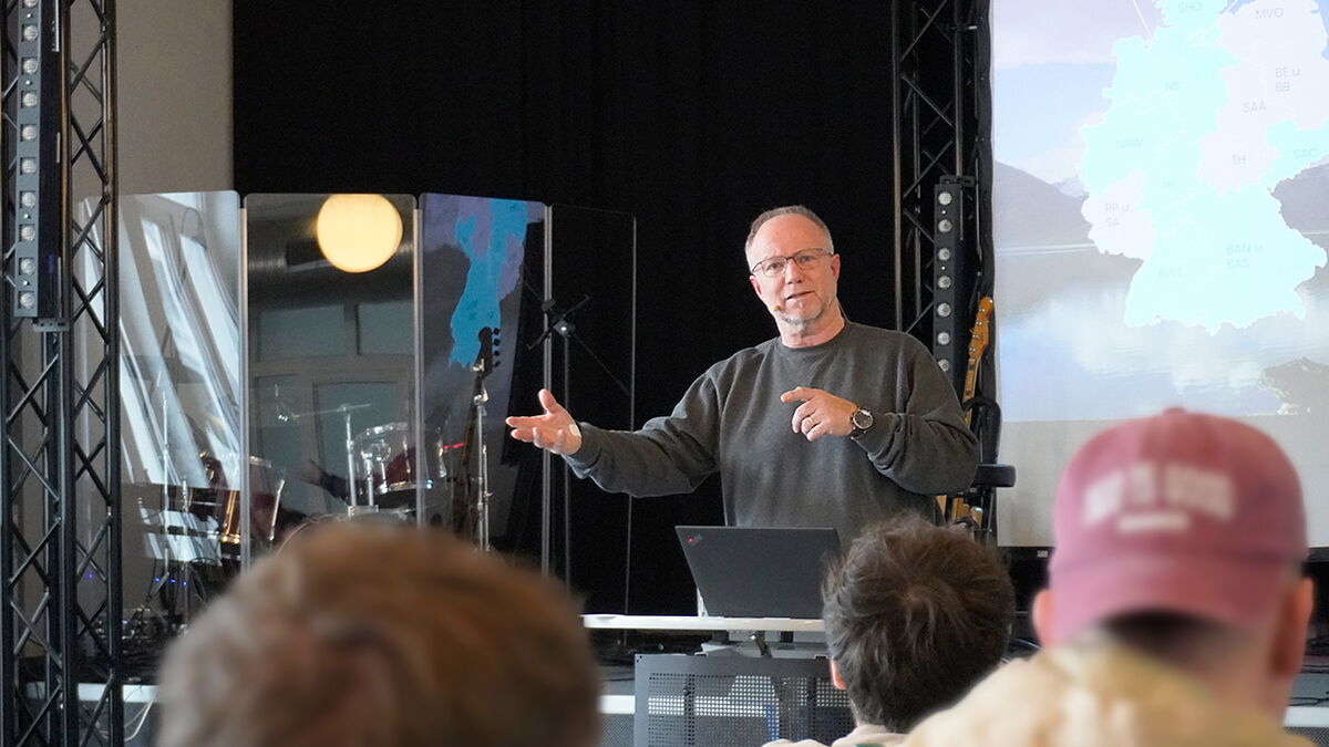 Mann steht vor einer Gruppe und spricht mit Gestik zu den Zuhörenden; im Hintergrund eine Präsentationsfolie und Bühnenausstattung mit Instrumenten und Technik.