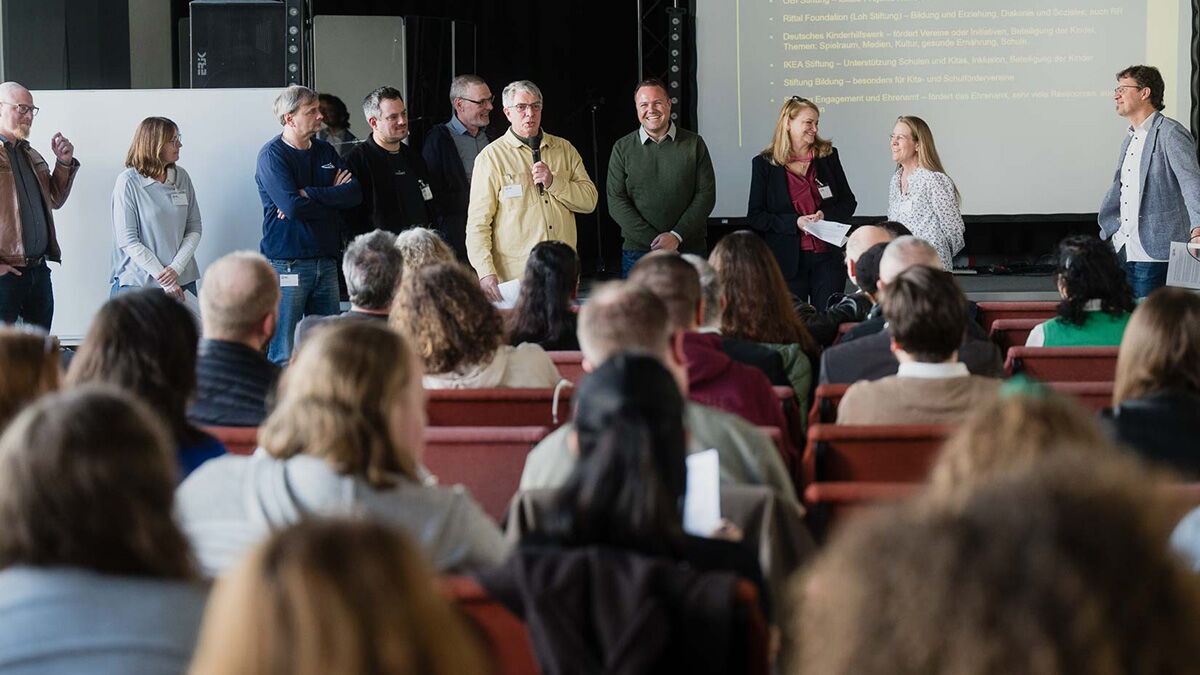 Joachim Kristahn steht mit Mikrofon in der Mitte einer Gruppe von Referentinnen und Referenten und stellt vor Publikum die Workshopthemen des Forumstags in Erzhausen vor.