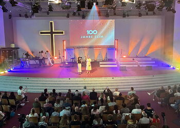 Blick in den Saal der Arche Hamburg-Stellingen beim Festgottesdienst zum 100-jährigen Jubiläum der ELIM Kirche Hamburg. Auf der Bühne steht vor einem großen Kreuz die Projektion „100 Jahre ELIM“, davor sitzen zahlreiche Gäste.