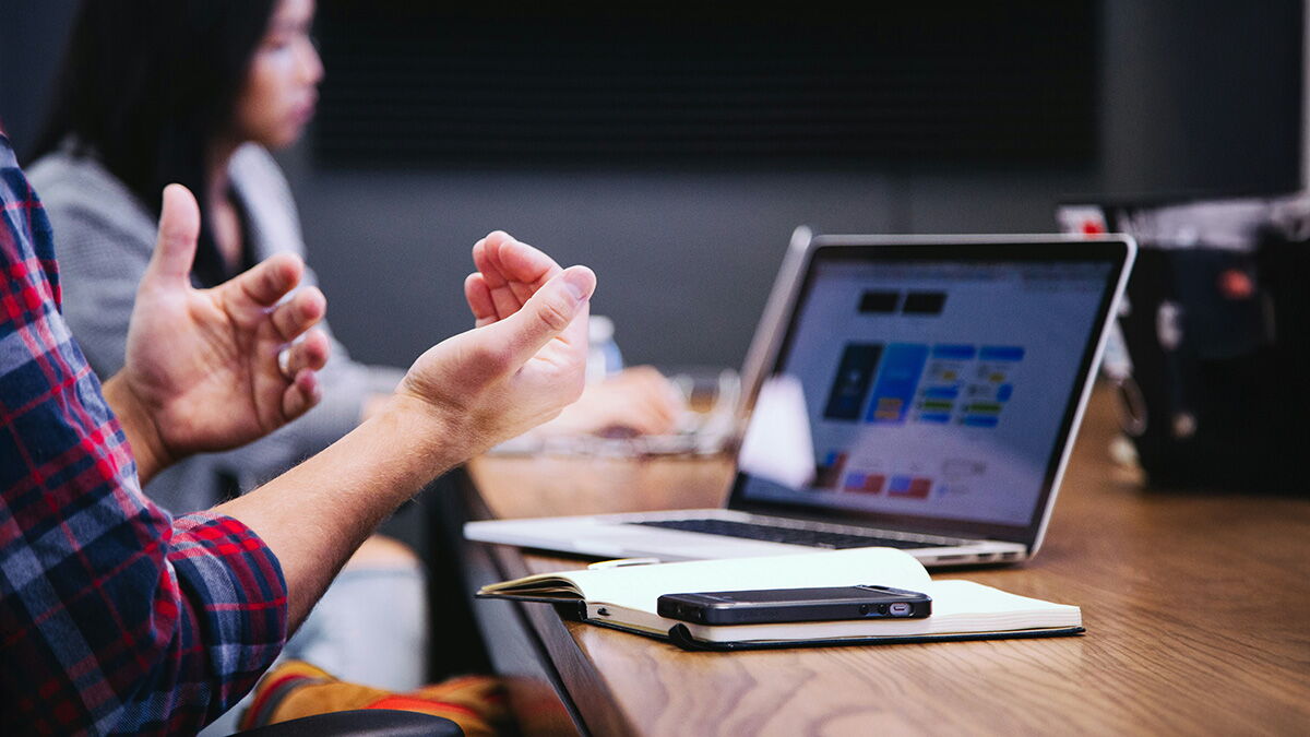 Mann vor einem aufgeklapptem Notebook mit einem Notitzbuch auf dem Tisch und einer sitzenden Frau im Hintergrund