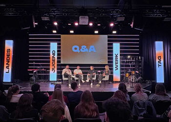 Mehrere Personen sitzen auf einer Bühne bei einem Q&A-Podium, davor Publikum; große Leinwand und beleuchtete Banner mit „Jugend“, „Werk“ und „Tag“