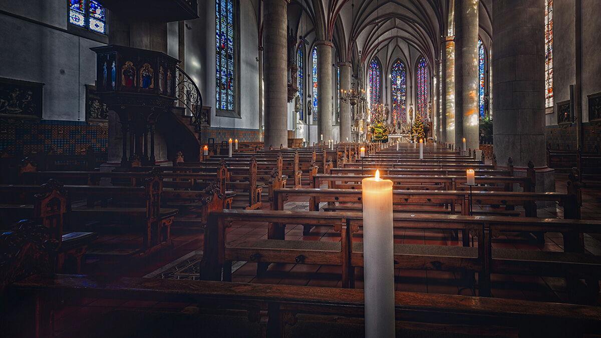 Innenraum einer großen Kirche mit hohen Gewölben, bunten Glasfenstern und Holzbänken; im Vordergrund brennt eine einzelne Kerze, der Raum wirkt ruhig und lichtdurchflutet.