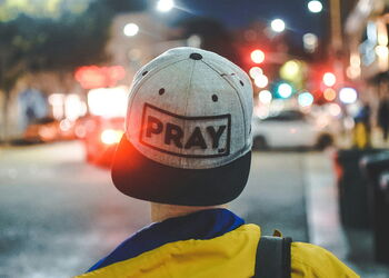 Person mit einer grauen Kappe mit der Aufschrift PRAY blickt auf eine nächtliche, beleuchtete Straße.