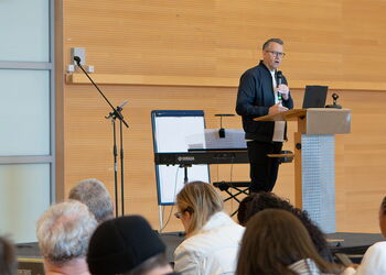 Ein Mann steht an einem Rednerpult und hält einen Vortrag vor einem Publikum in einem großen Konferenzsaal.