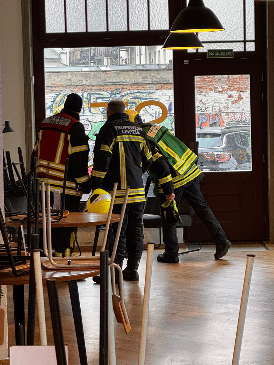 Drei Männer in Feuerwehrkleidung begutachten einen Schadensort in einem Café.