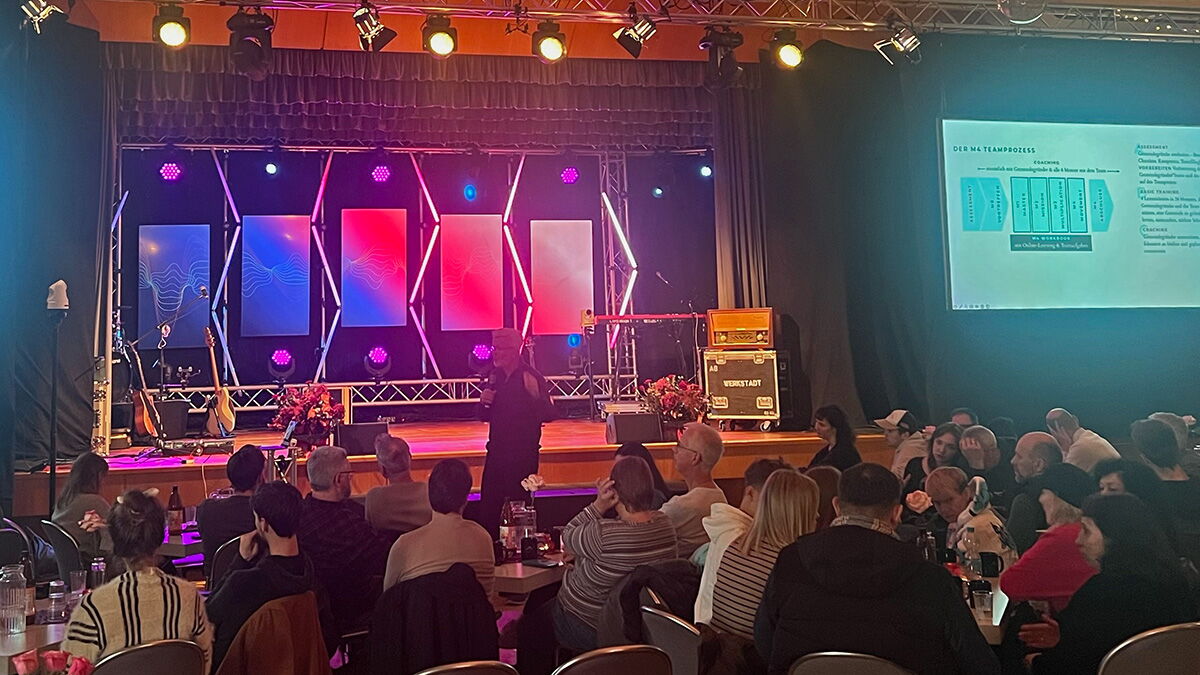 Zuhörer sitzen in Tischgruppen bei einem Workshop - der Referent steht vor der Bühne.