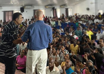 Zwei Menschen stehen auf einer Bühne mit dem Rücken zum Fotografen, davor Menschen, die die Hände heben und beten.