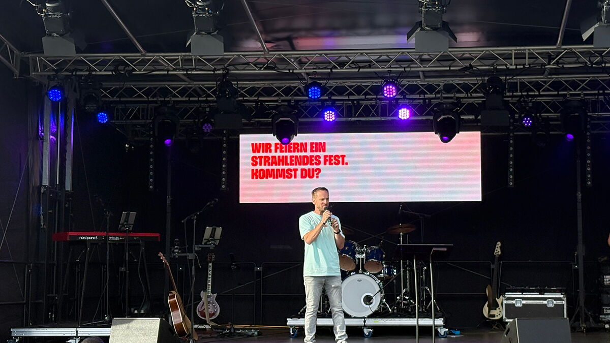 Martin Kopp spricht mit einem Mikrofon auf der HELLER-Bühne auf dem Darmstädter Friedensplatz. Hinter ihm steht auf der LED-Wand: „Wir feiern ein strahlendes Fest. Kommst du?“