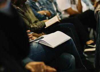 Teilnehmende eines Seminars sitzen in Reihen; im Vordergrund macht eine Person Notizen in einem aufgeschlagenen Notizbuch.