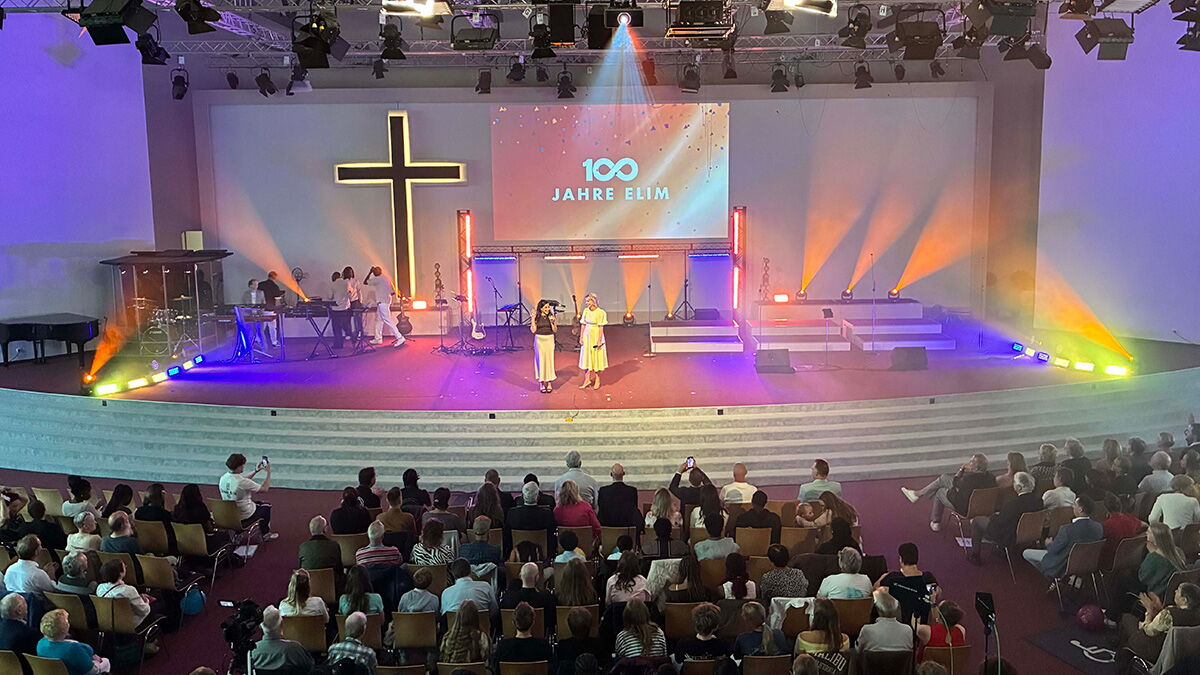 Blick in den Saal der Arche Hamburg-Stellingen beim Festgottesdienst zum 100-jährigen Jubiläum der ELIM Kirche Hamburg. Auf der Bühne steht vor einem großen Kreuz die Projektion „100 Jahre ELIM“, davor sitzen zahlreiche Gäste.