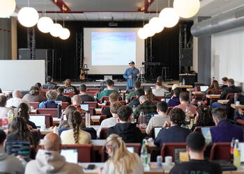 Großer Seminarraum mit vielen sitzenden Teilnehmerinnen und Teilnehmern an Tischen, die einem Referenten vor einer Leinwand zuhören; einige arbeiten mit Laptops, vorne ist eine Bühne mit Präsentation und Musikinstrumenten.