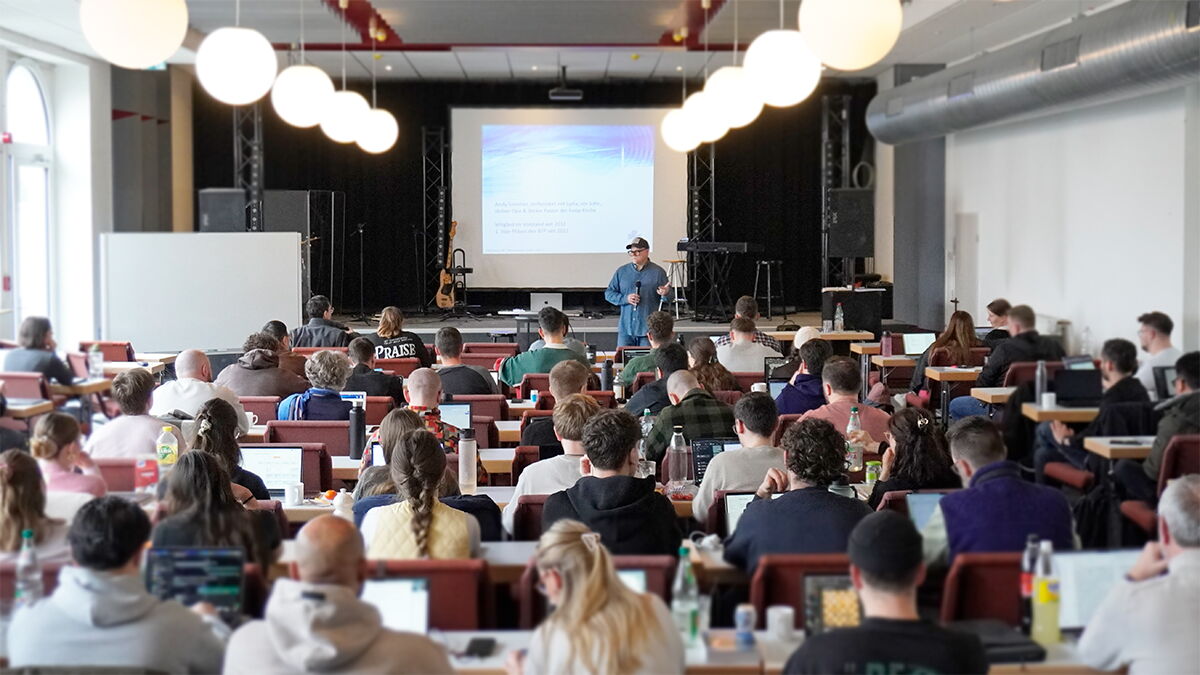 Großer Seminarraum mit vielen sitzenden Teilnehmerinnen und Teilnehmern an Tischen, die einem Referenten vor einer Leinwand zuhören; einige arbeiten mit Laptops, vorne ist eine Bühne mit Präsentation und Musikinstrumenten.