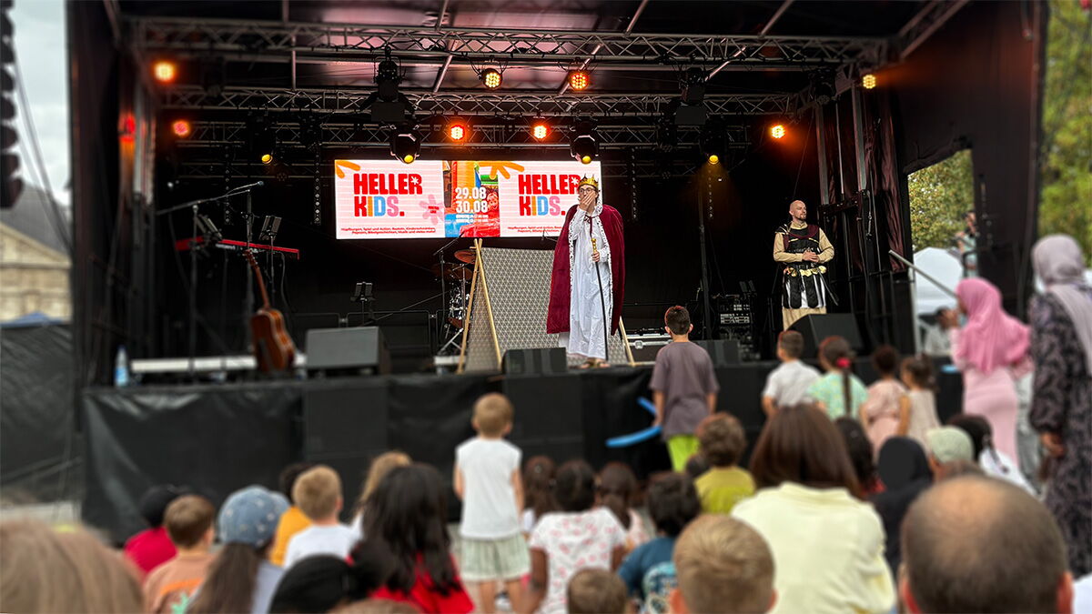 Kinder und Erwachsene verfolgen vor der HELLER-Bühne auf dem Friedensplatz ein Theaterstück. Zwei kostümierte Darsteller stehen auf der Bühne.