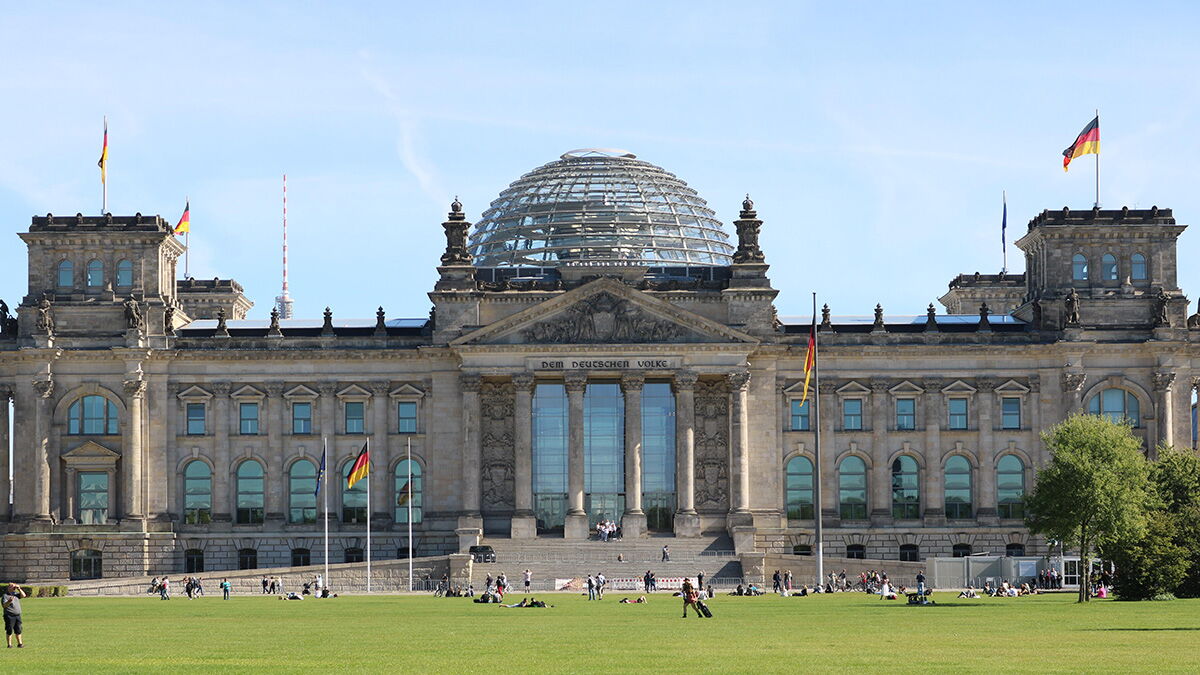 Reichstagsgebäude in Berlin mit Glaskuppel und Deutschlandflaggen vor blauem Himmel.