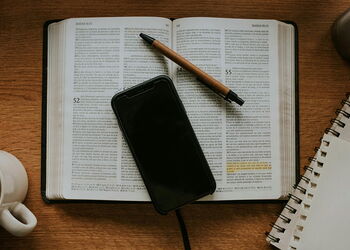 Eine aufgeschlagene Bibel und ein Smartphone liegen neben einem Kaffeebcher und Schreibheft auf einem Tisch.