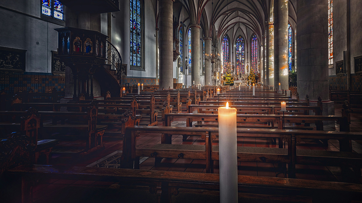 Innenraum einer großen Kirche mit hohen Gewölben, bunten Glasfenstern und Holzbänken; im Vordergrund brennt eine einzelne Kerze, der Raum wirkt ruhig und lichtdurchflutet.