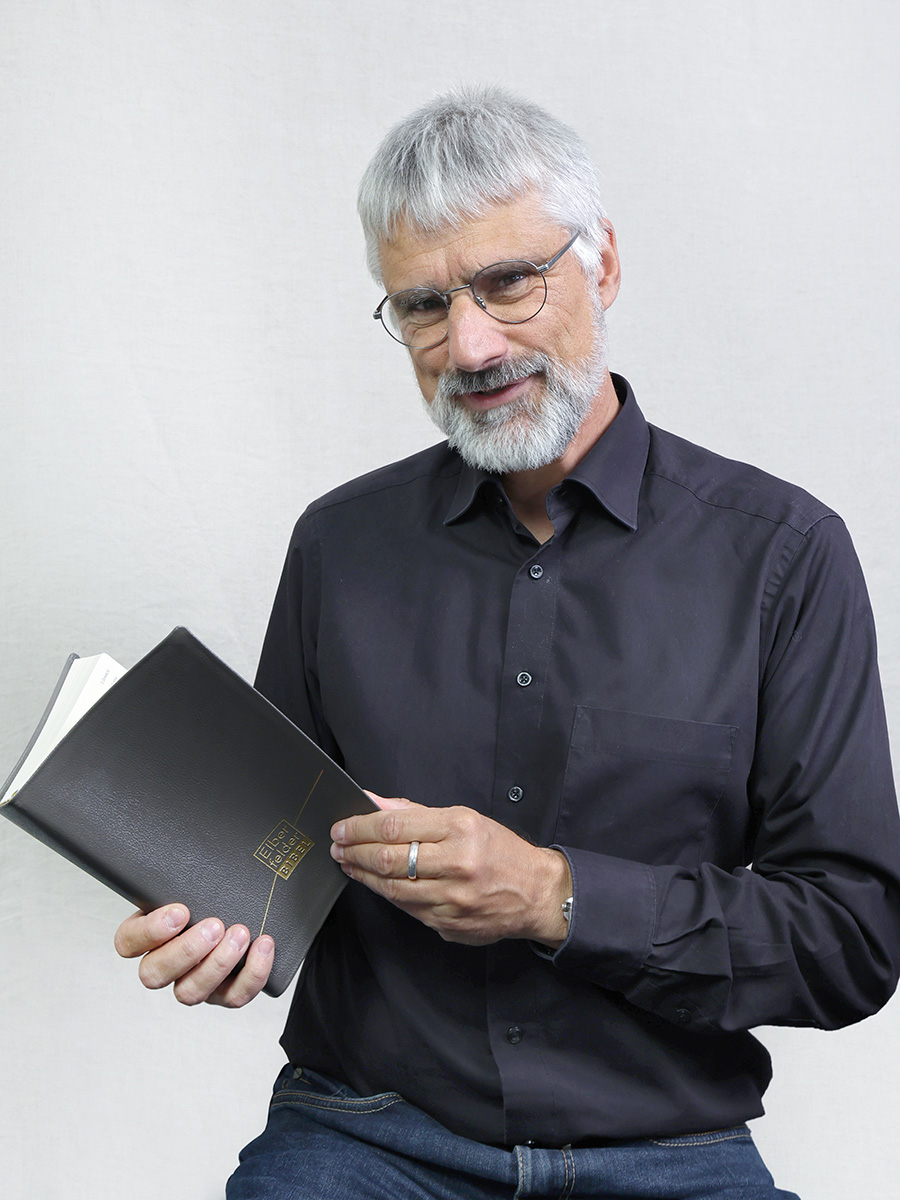 An Mann hält eine Bibel in der Hand und schaut freundlich in die Kamera.